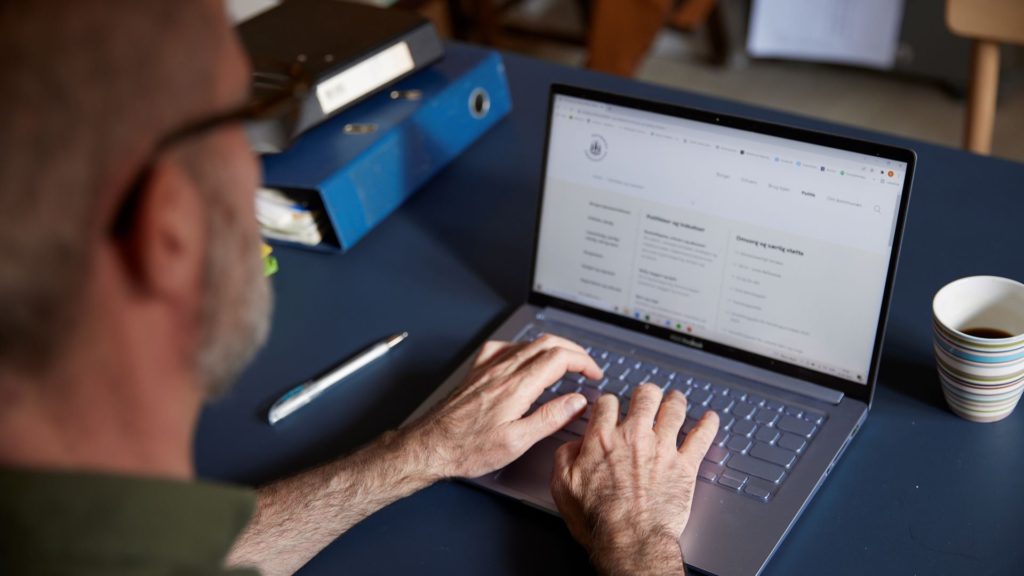 person arbejder på en bærbar computer ved et bord med en pen og mapper. Skærmen viser et websted med tekst og menuer.