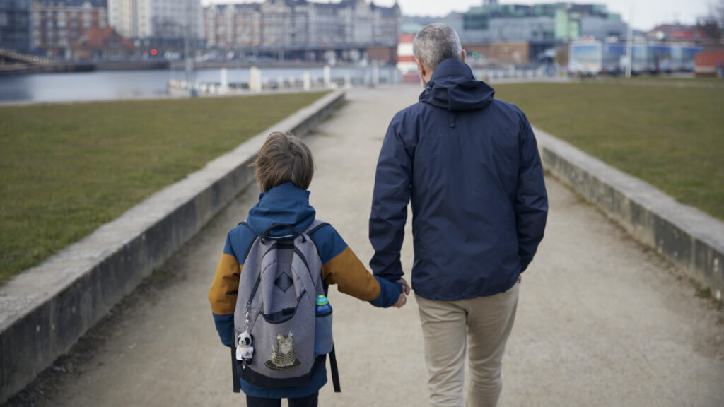 voksen og et barn, der holder i hånd, går på en sti udendørs. Barnet har rygsæk på og byen ses i baggrunden.