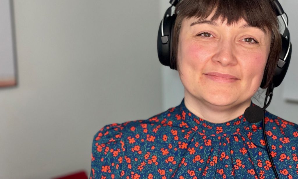 kvinde med korte brune hår og hovedtelefoner smiler, iført en blå bluse med røde blomster.