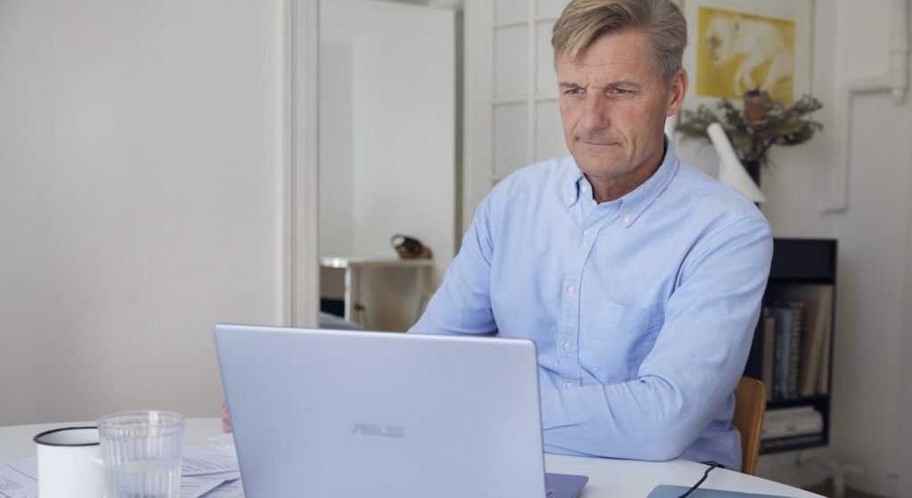 Mand sidder ved et skrivebord og arbejder på en bærbar computer i et lyst rum.
