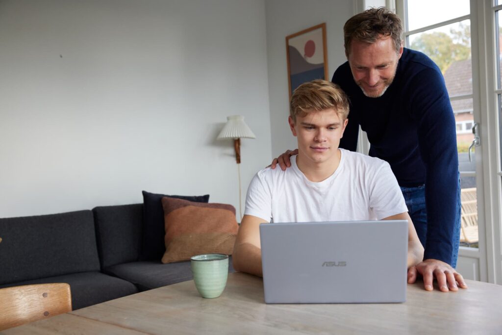 mænd sidder ved et bord med en bærbar computer. Den ene mand kigger på skærmen, mens den anden står bag ham og smiler.