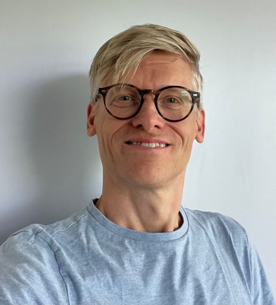Mand blondhåret mand med briller og blå t-shirt smiler foran en neutral, lys baggrund.