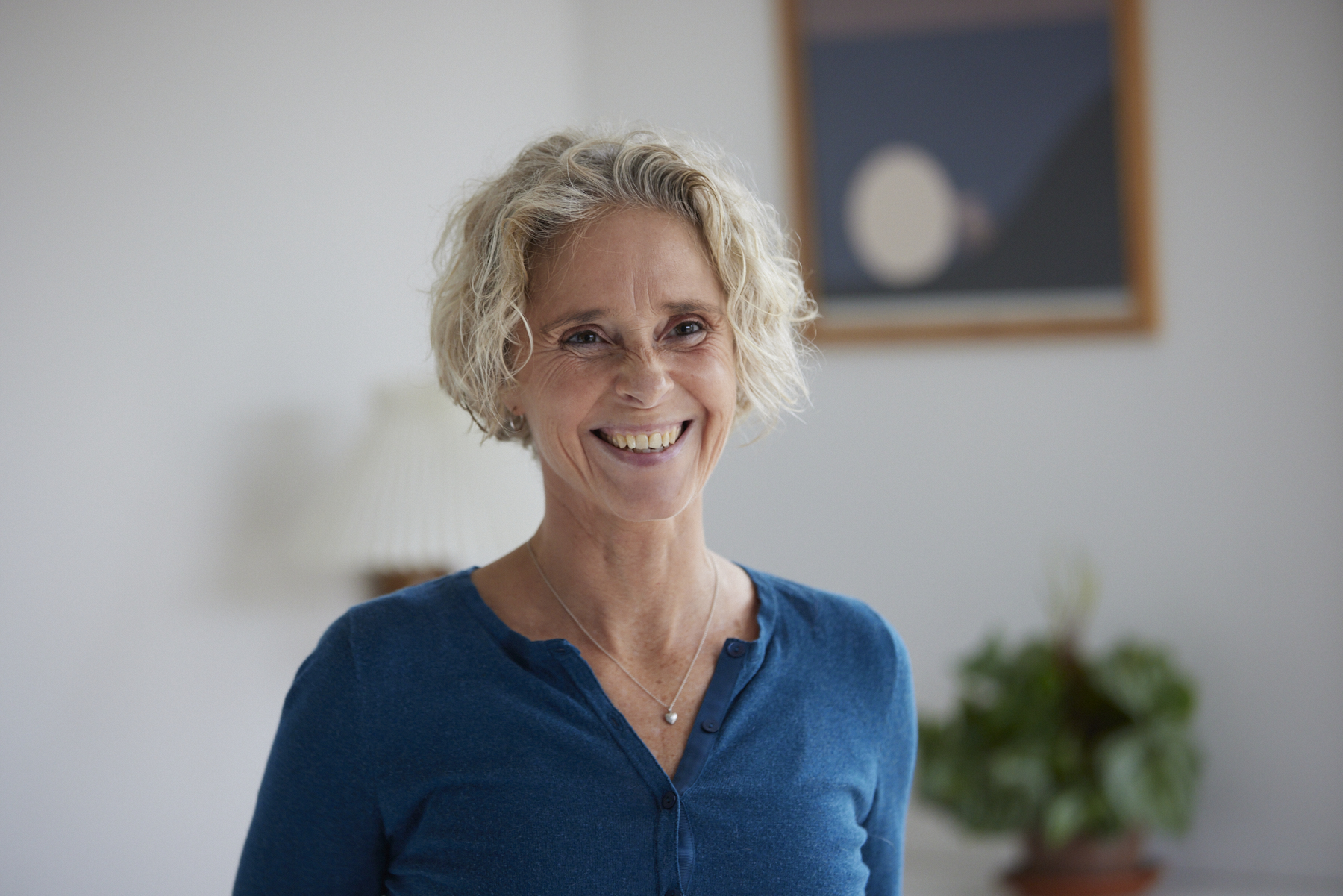 smilende, lyshåret kvinde i en blå trøje står indendørs med planter og et maleri i baggrunden.