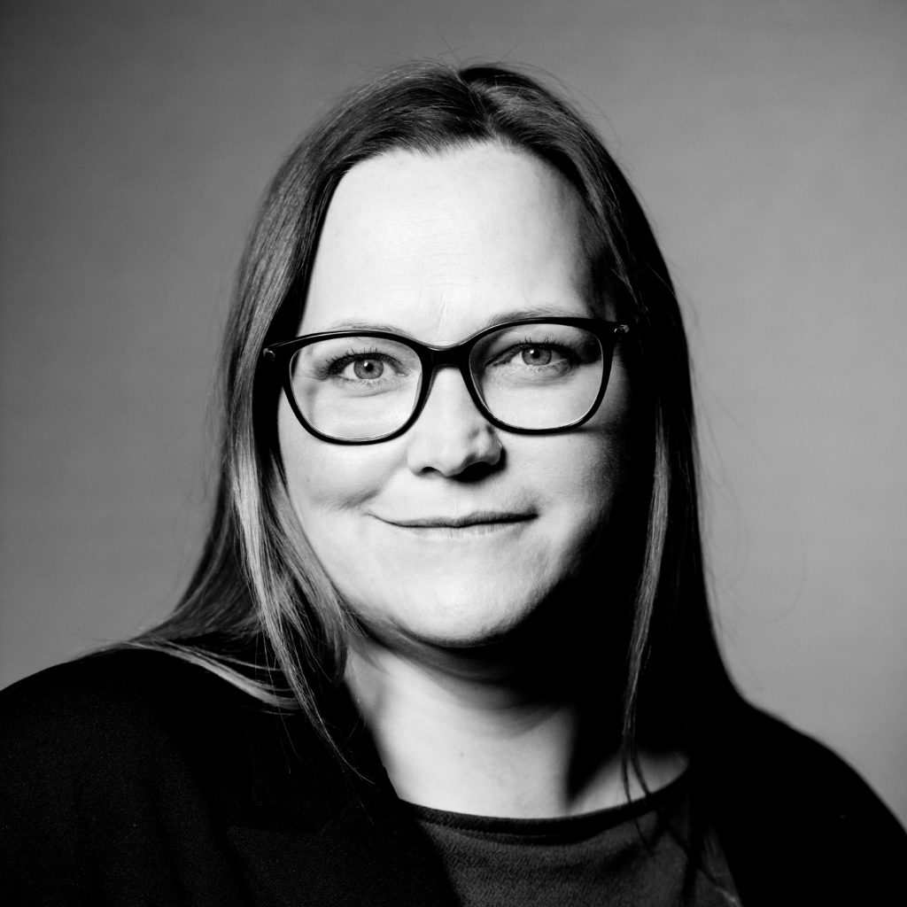 Sort-hvid portræt af kvinde med langt hår og briller, iført mørkt tøj, ser direkte ind i kameraet med et neutralt udtryk.
