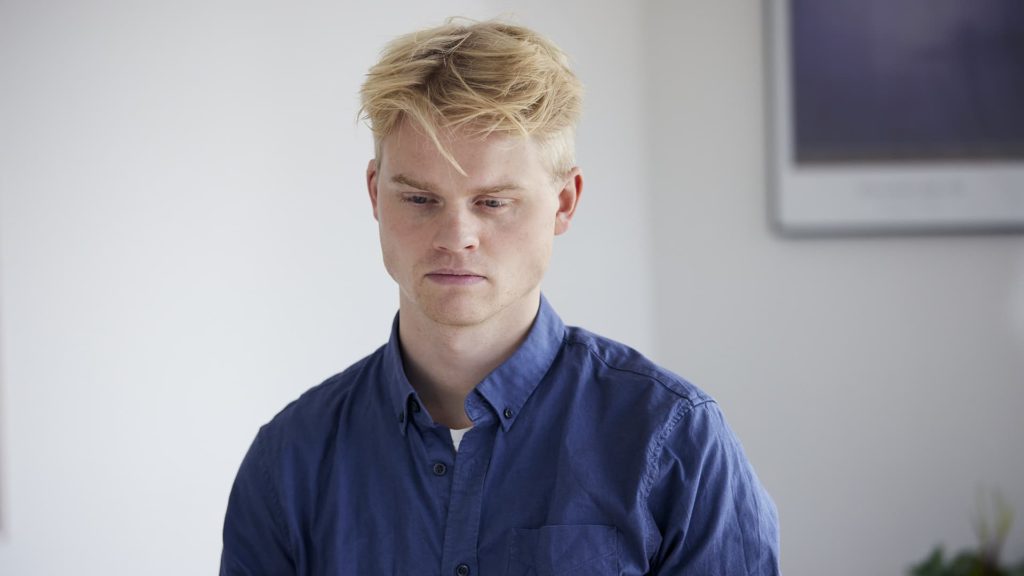 Ung mand med blondt hår iført en blå skjorte kigger nedad med et neutralt udtryk i et lyst rum.