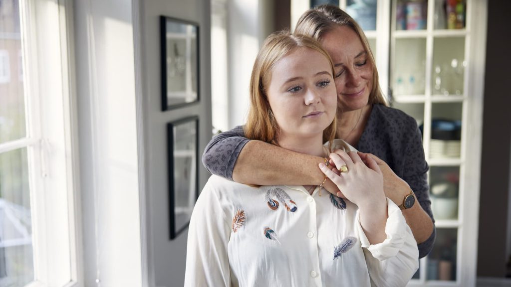 kvinde krammer en ung kvinde bagfra i et hjemligt miljø, de ser rolige og tætte ud.