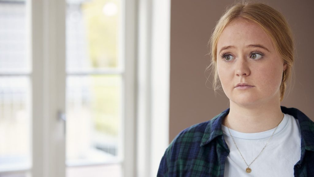 ung kvinde med blondt hår ser bekymret ud, mens hun står indendørs nær et vindue.