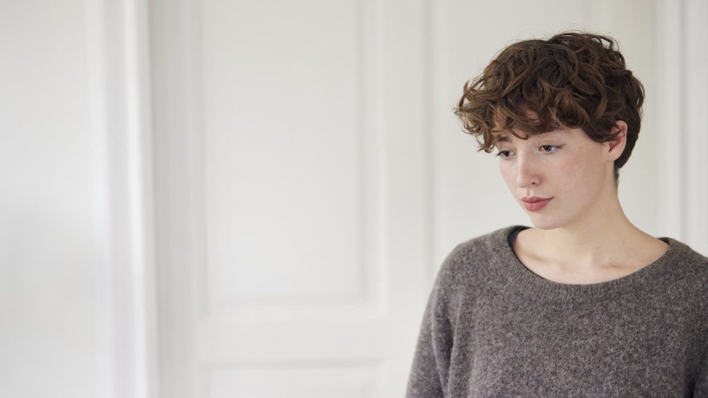 Person med kort, krøllet hår iført en grå sweater står foran en hvid væg og kigger nedad.