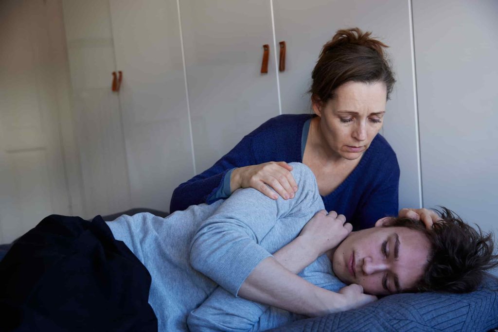 kvinde trøster en ung mand, der ligger i fosterstilling på en seng. Begge ser bekymrede ud.