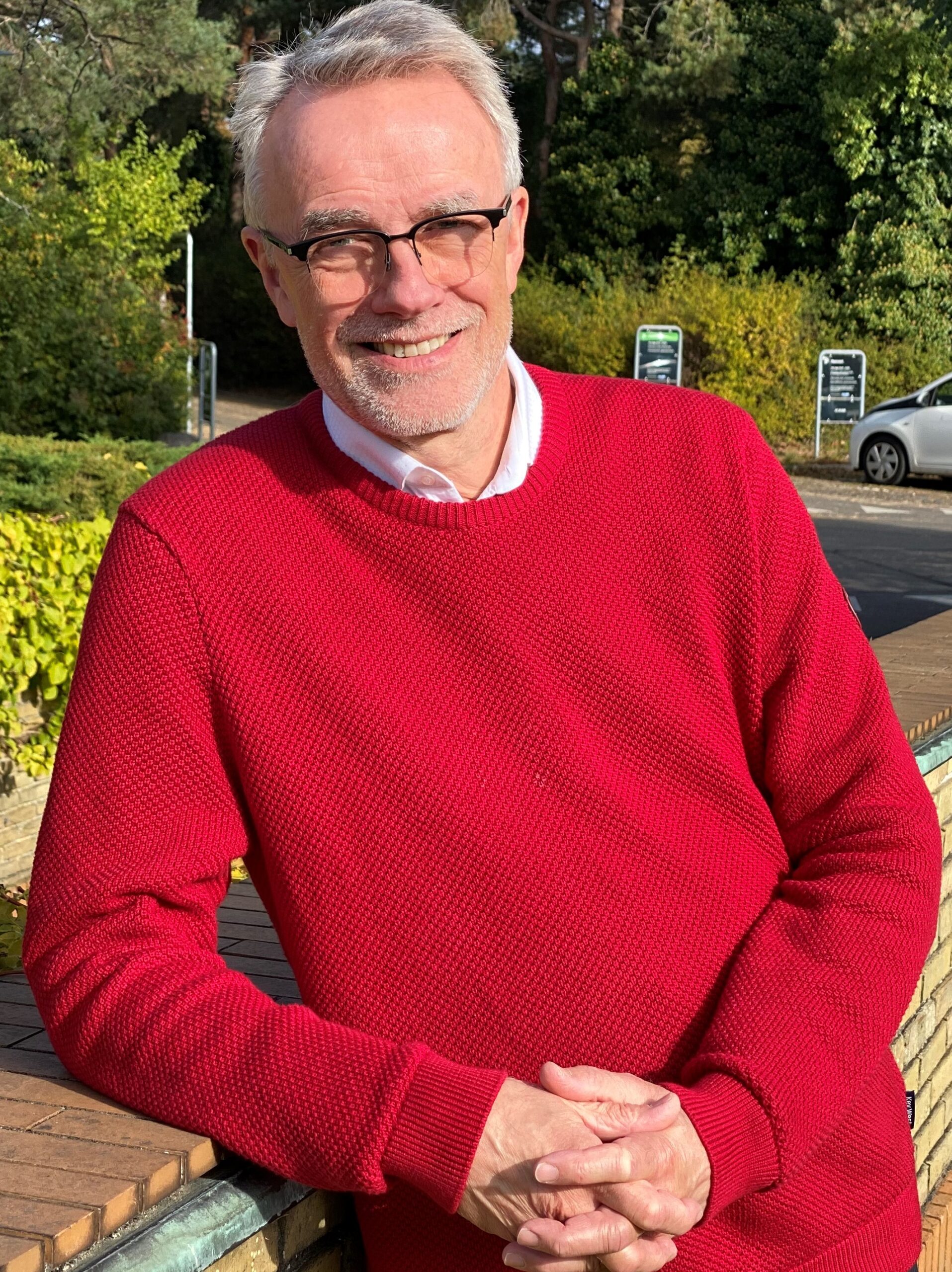 ældre mand med briller og rød sweater smiler, mens han står udenfor på en solrig dag med grønne træer i baggrunden.