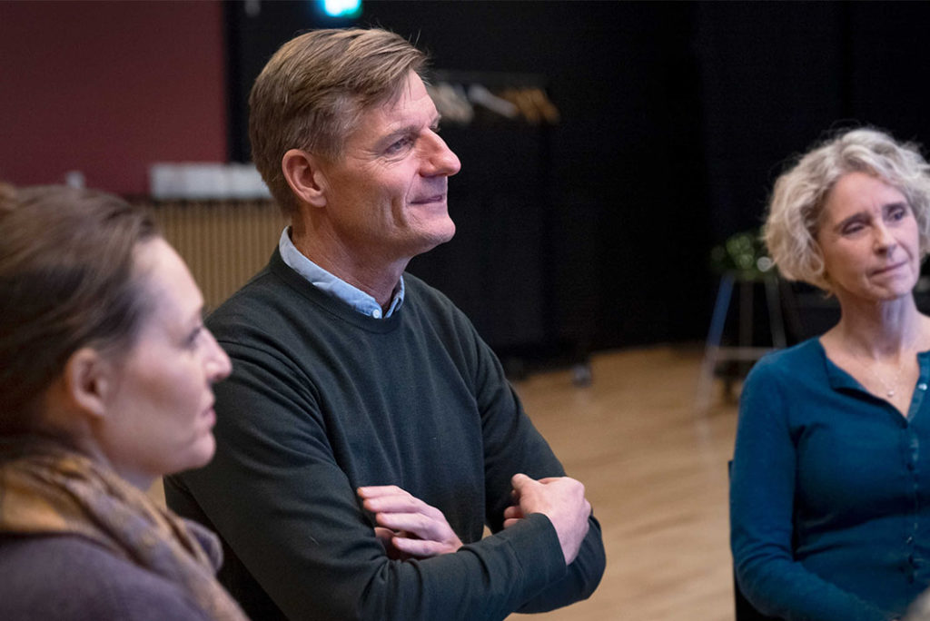 mand står med korslagte arme indendørs og smiler, mens han lytter opmærksomt til nogen uden for billedet. To andre personer er delvist synlige.