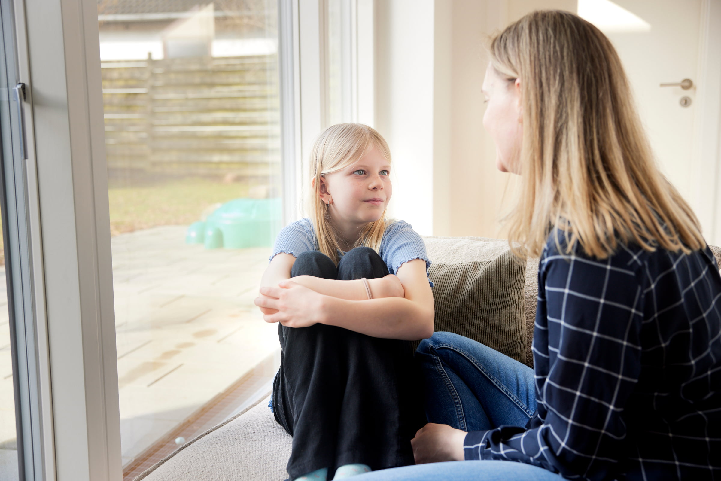 pige sidder med benene trukket op, mens hun taler med en kvinde indendørs ved et stort vindue.