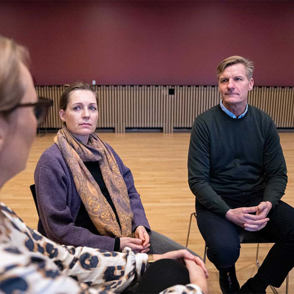 Tre personer sidder i en kreds og taler sammen i et stort rum med trægulv og rød væg.