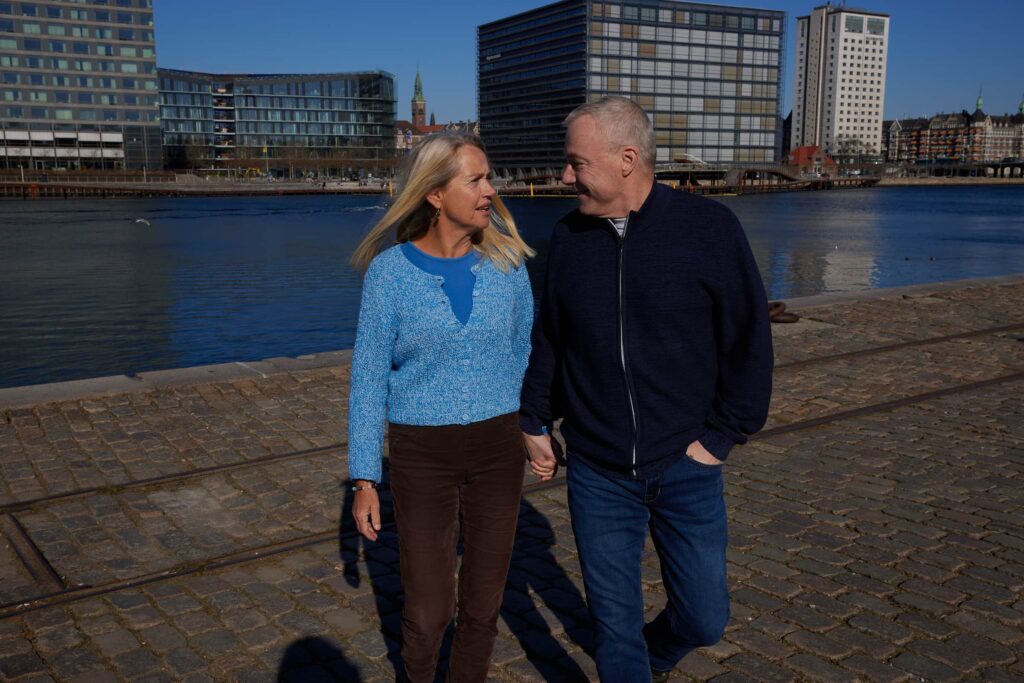 ældre par går hånd i hånd ved vandet, med moderne bygninger og klart vejr i baggrunden.