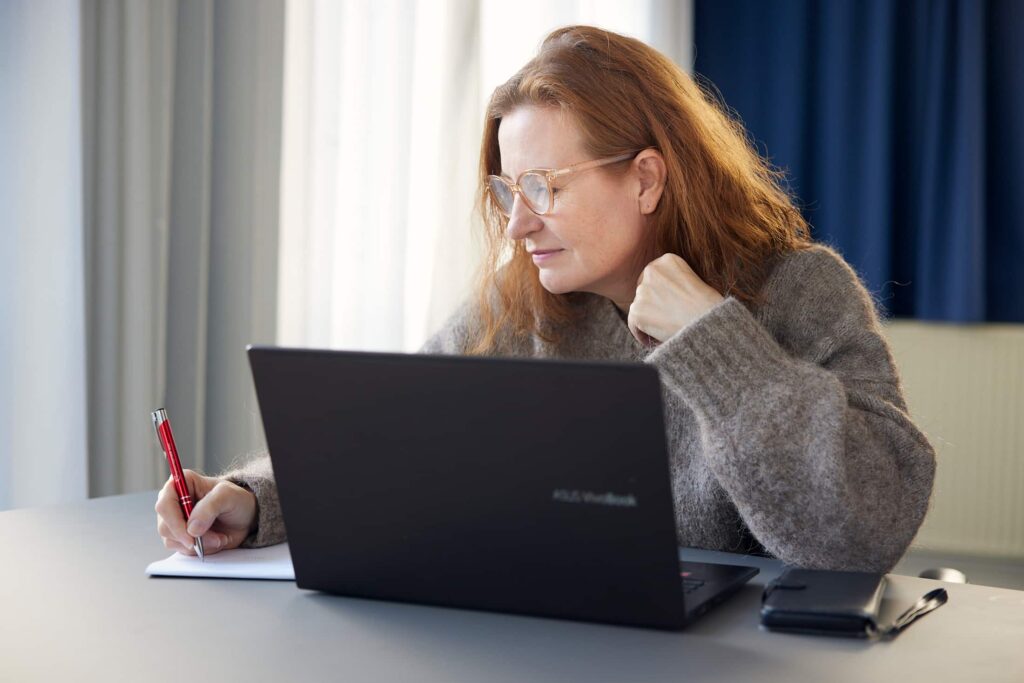 Kvinde med briller sidder ved skrivebord, arbejder på en bærbar computer og skriver på papir med en rød pen.