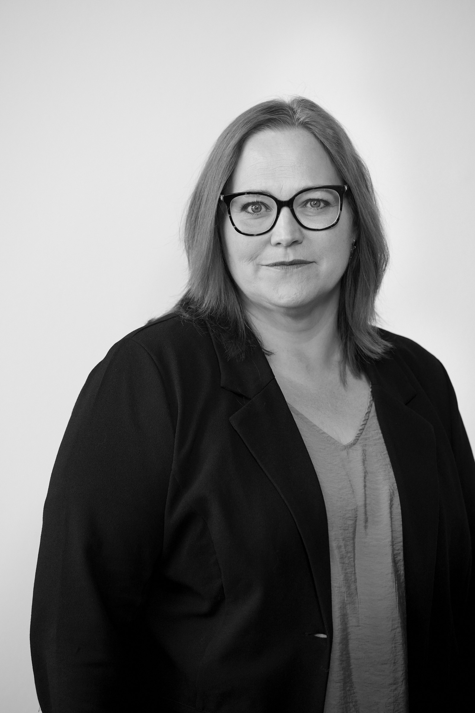 kvinde med skulderlangt hår og briller, iført blazer og bluse, står foran en lys baggrund. Billedet er i sort-hvid.