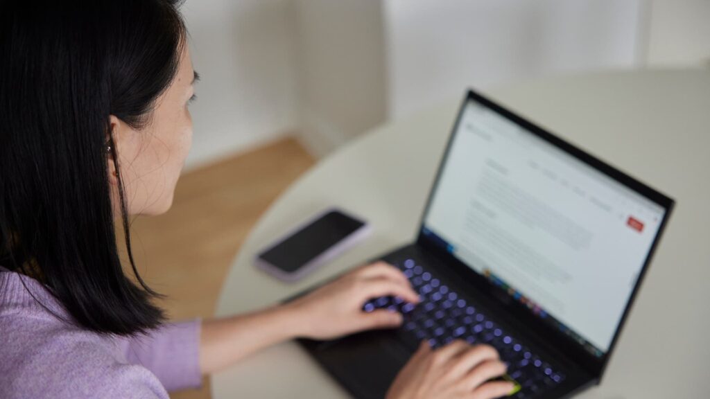 Person arbejder ved et rundt bord på en bærbar computer, med en smartphone liggende ved siden af.