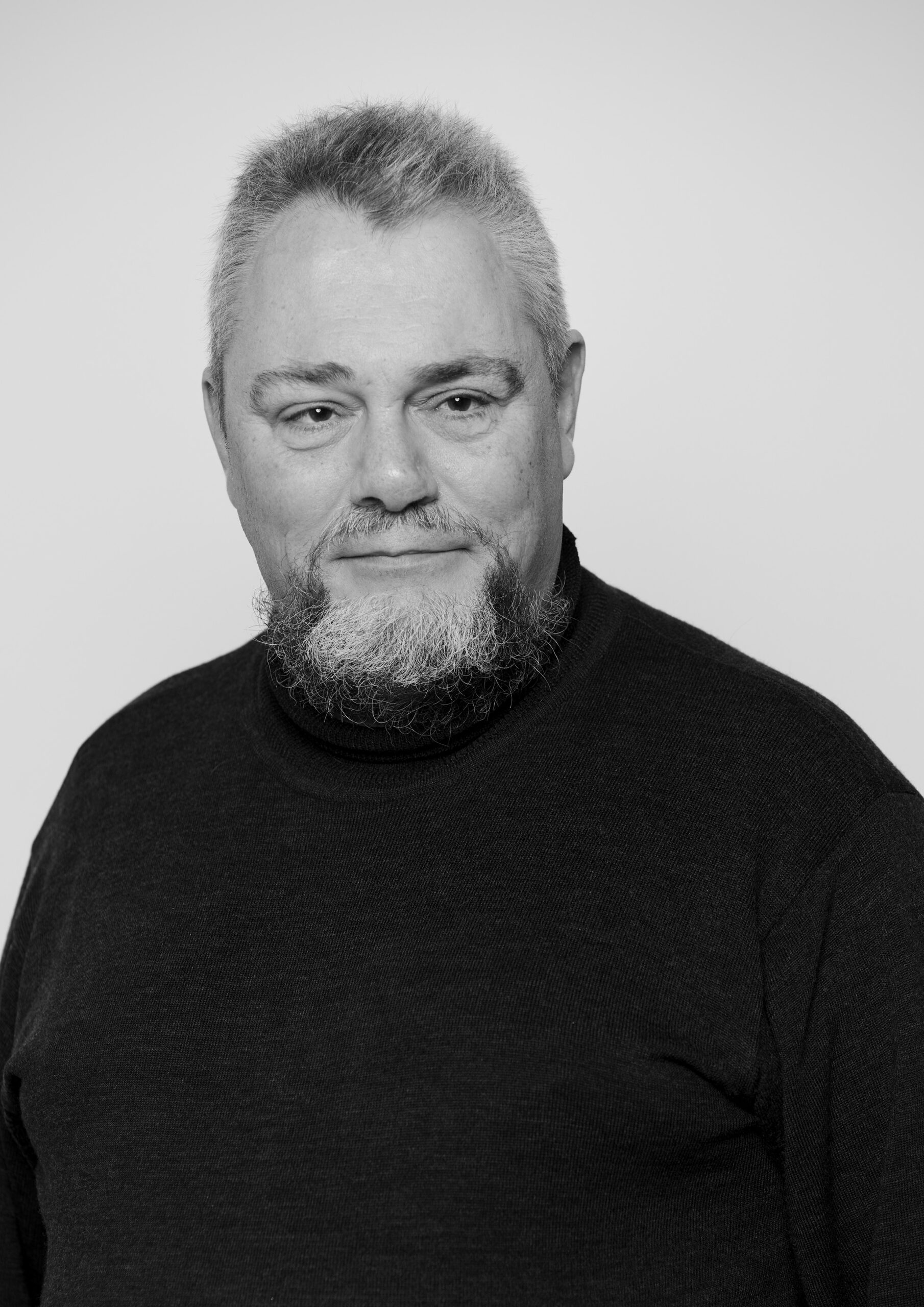 Sort-hvidt portræt af mand med kort hår og fuldskæg iført en mørk trøje. Han kigger roligt ind i kameraet med et svagt smil.