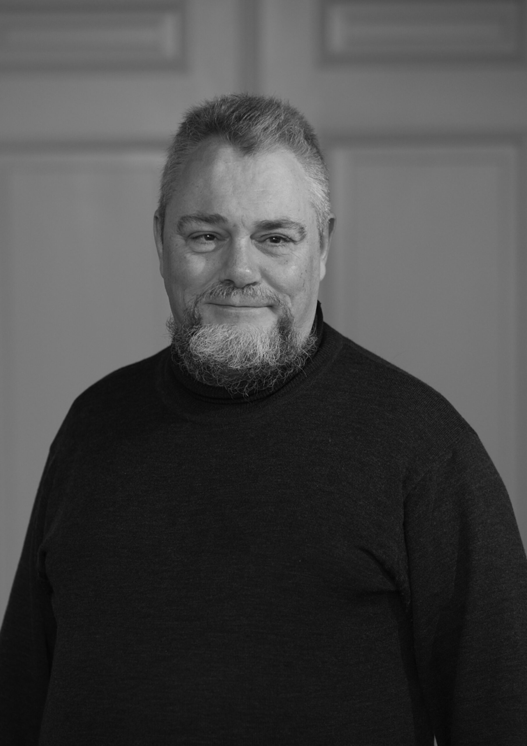 Mand portrætteret i sort-hvid, iført mørk trøje, smilende let med kort gråt hår og fuldskæg. Ensfarvet baggrund.