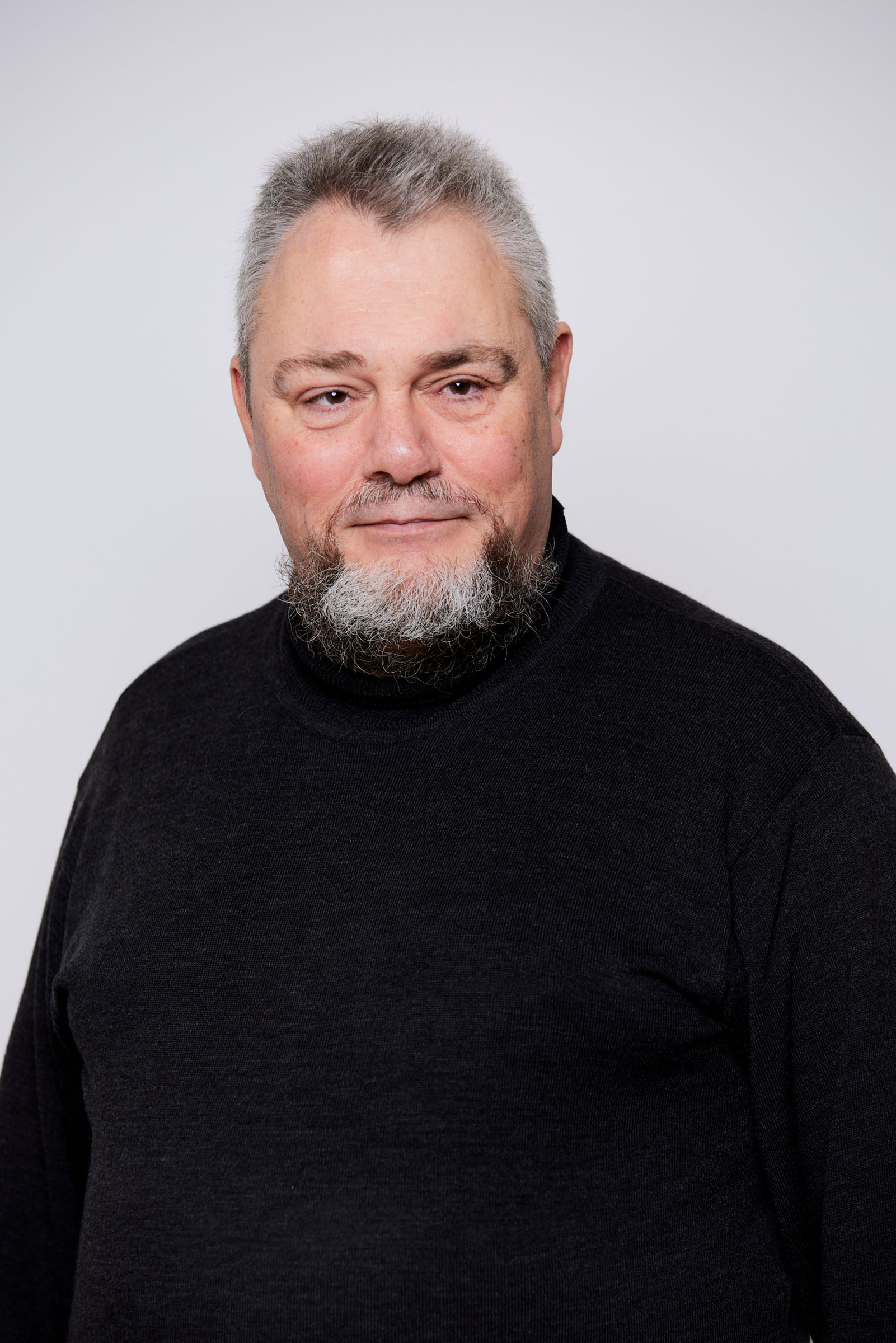 ældre mand med gråt hår og skæg iført en sort sweater, står foran en lyst baggrund og kigger roligt ind i kameraet.