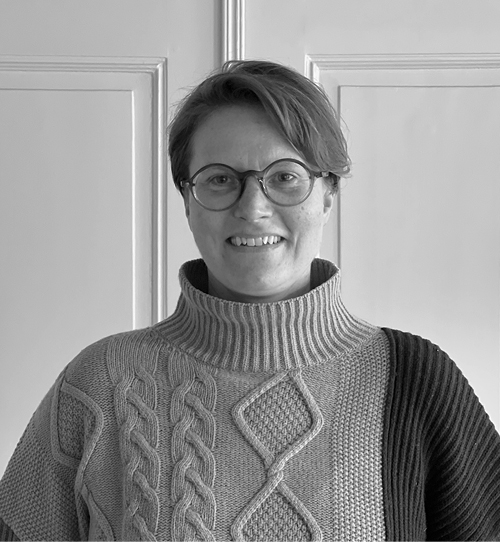 person med kort hår og briller smiler, iført en tyk striktrøje med forskellige mønstre, foran en lys baggrund.