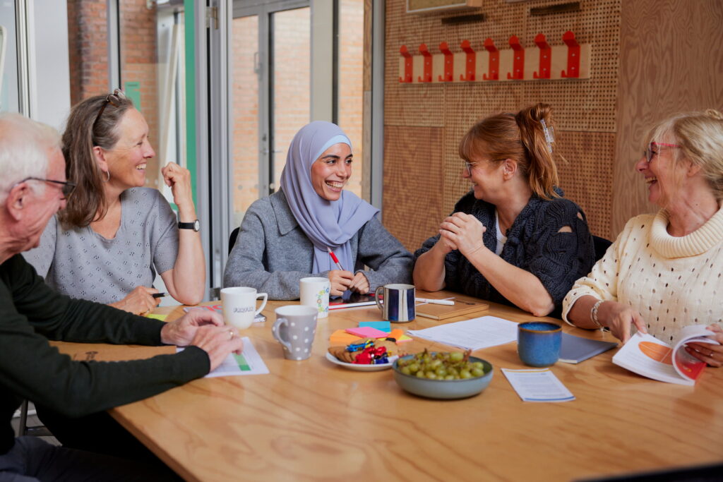 Fire kvinder sidder rundt om et bord og smiler, omgivet af kaffe, snacks og papirer, i et lyst og afslappet mødelokale.