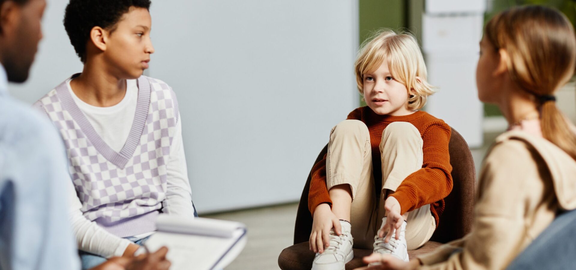 barn med blondt hår sidder på en stol med benene oppe, iført brun trøje og beige bukser, taler med en anden person indendørs.