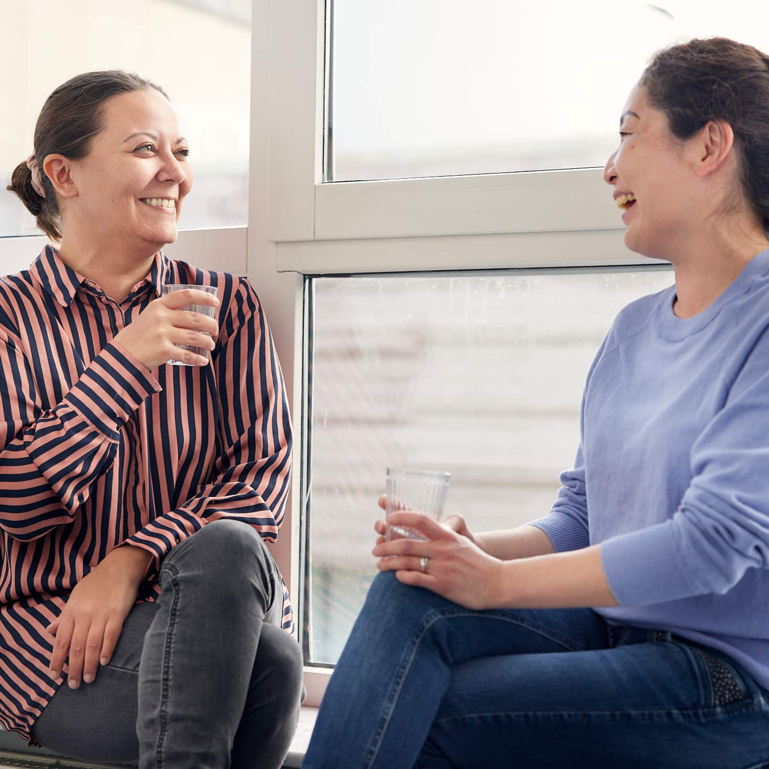 kvinder sidder ved et vindue og taler sammen, mens de smiler og holder drikkeglas.