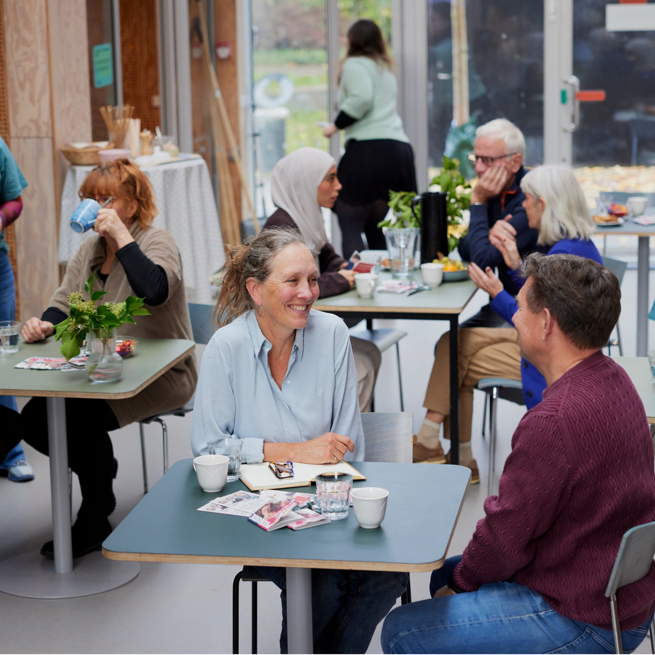 Folk sidder ved borde på en café, drikker kaffe, taler sammen og nyder en afslappet atmosfære.