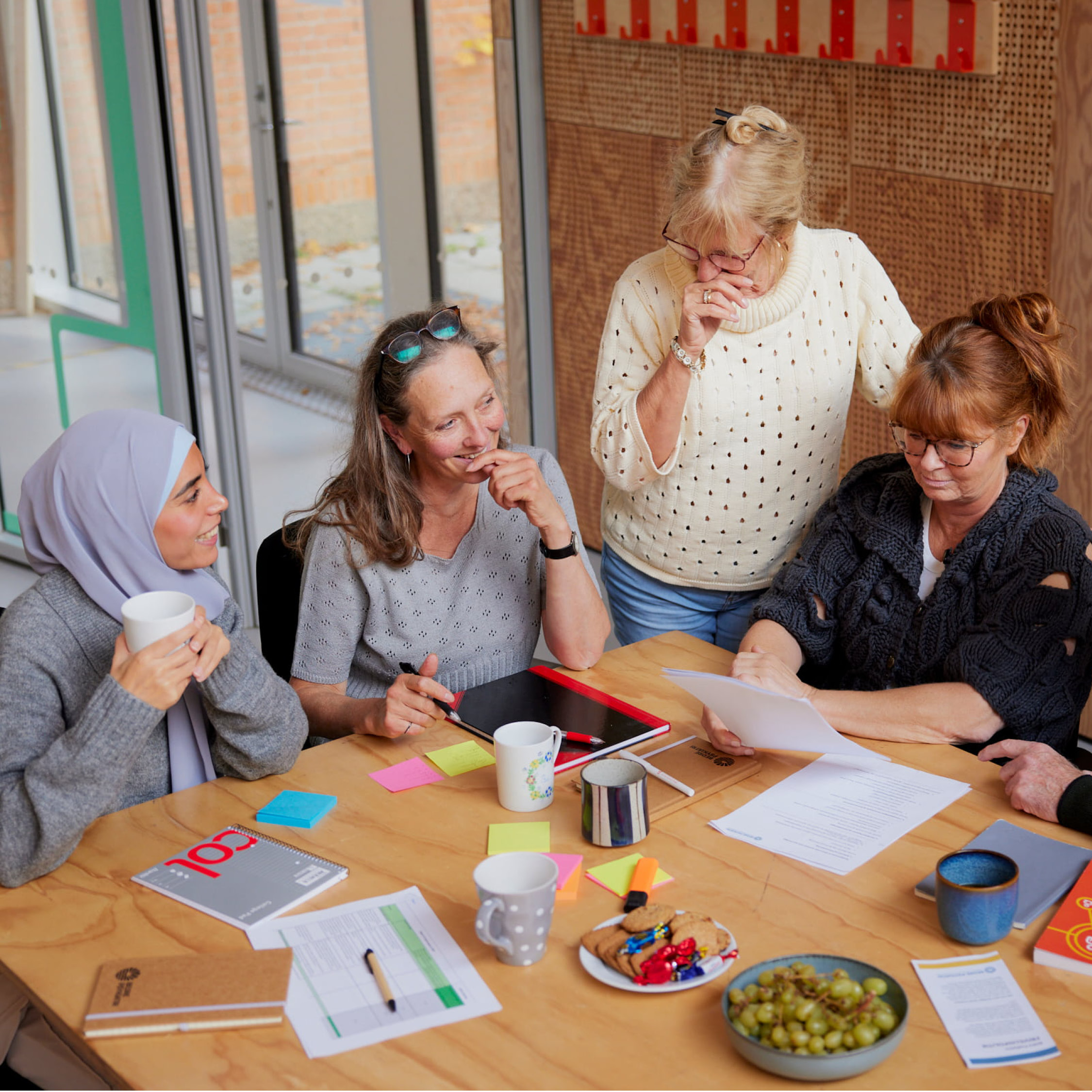 Fem kvinder sidder omkring et bord med dokumenter, noter, kaffe og snacks, engageret i en samtale eller gruppearbejde i et lyst lokale.