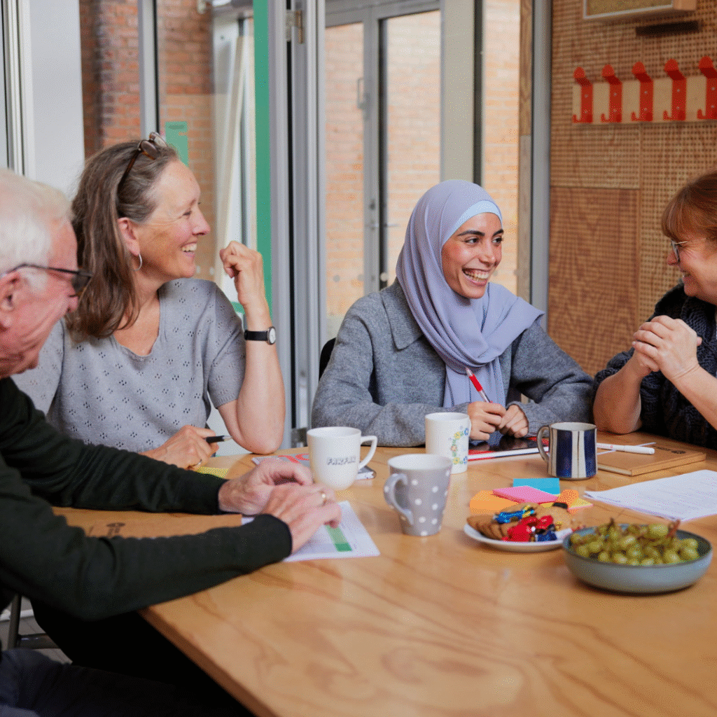 Fire personer sidder rundt om et bord, smiler og snakker sammen. Der er papir, kopper og snacks på bordet, og stemningen virker venlig og afslappet.