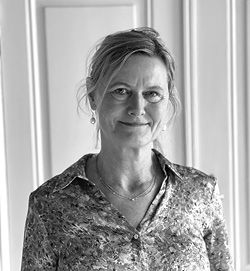 Sort-håret kvinde står foran en hvid væg iført mønstret skjorte, ser ind i kameraet og smiler let. Billedet er i sort-hvid.