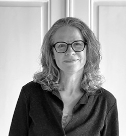 Sort-hvidt portræt af en kvinde med skulderlangt hår og briller, iført en mørk skjorte, stående foran en lys baggrund.