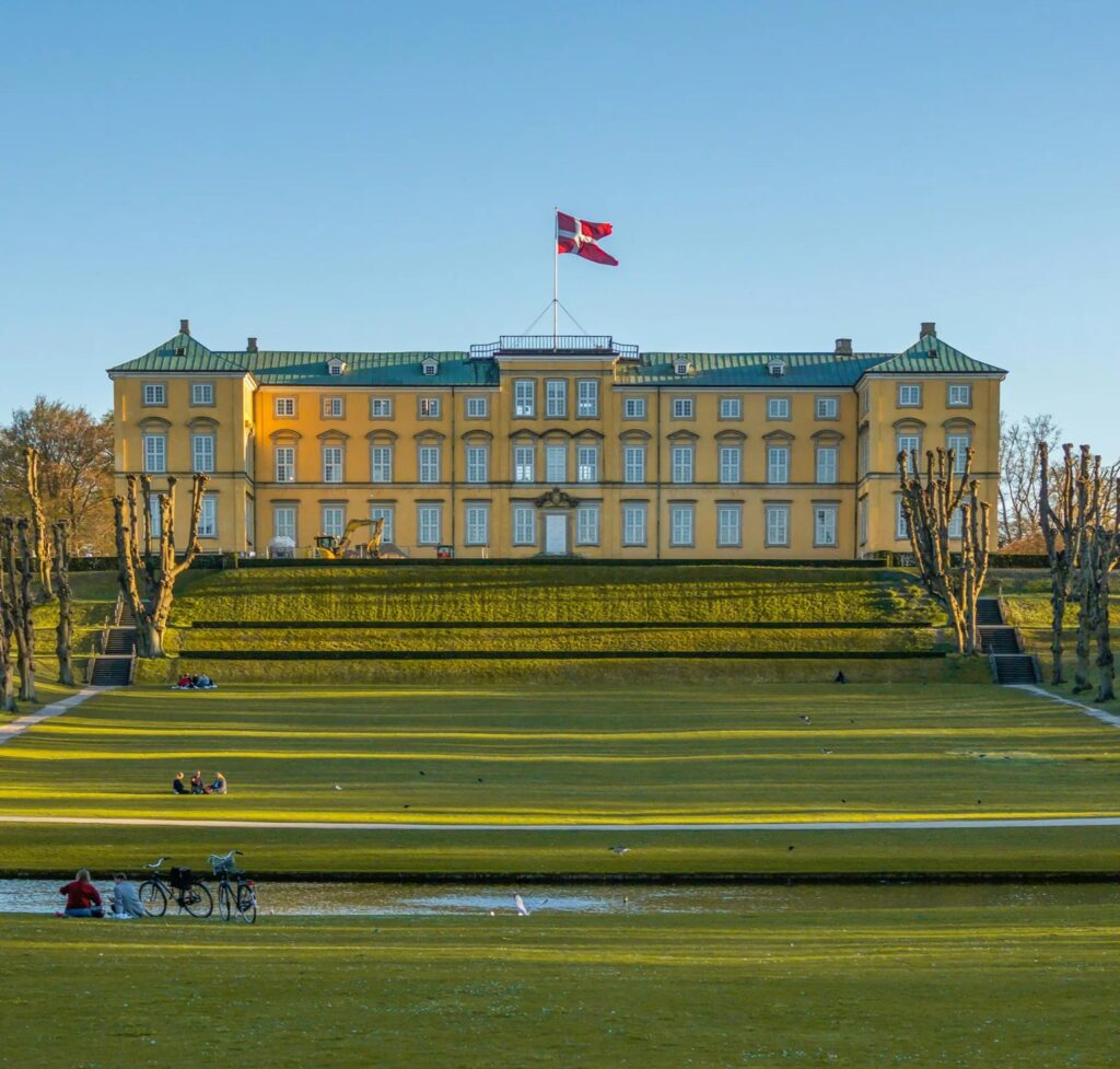 Gult slot med grønne tage og dansk flag, set fra en stor plæne med folk der slapper af i solen.