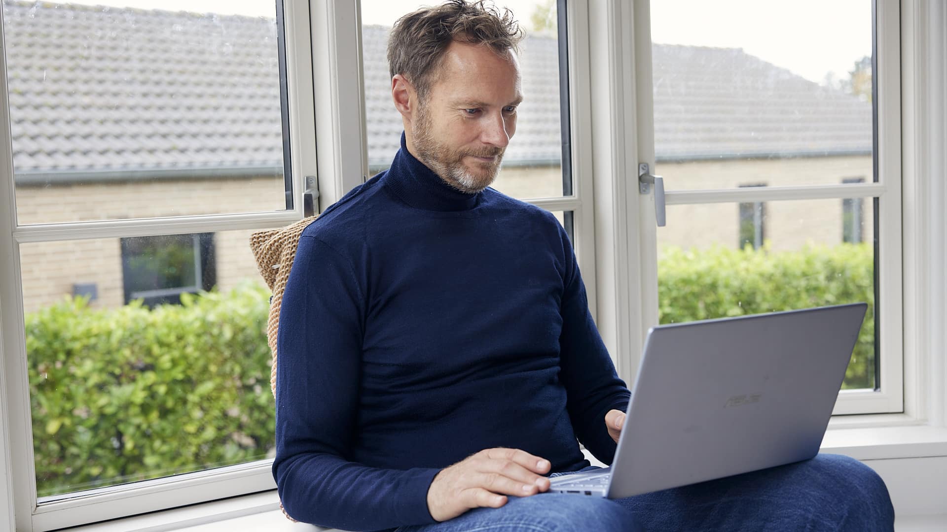 mand iført mørkeblå rullekrave sidder ved et vindue og arbejder på en bærbar computer, med grønne planter udenfor.