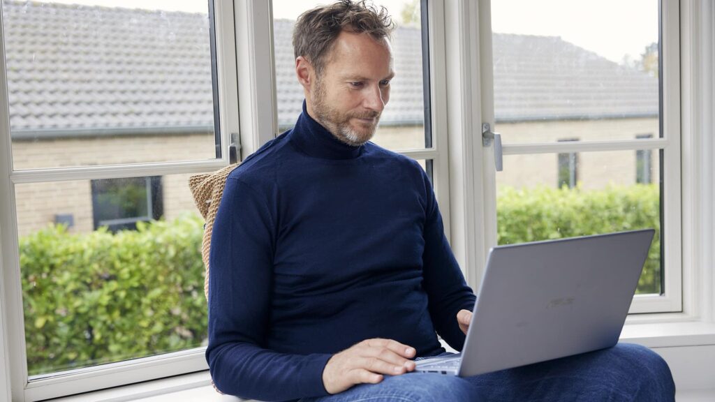 mand iført mørkeblå rullekrave sidder ved et vindue og arbejder på en bærbar computer, med grønne planter udenfor.
