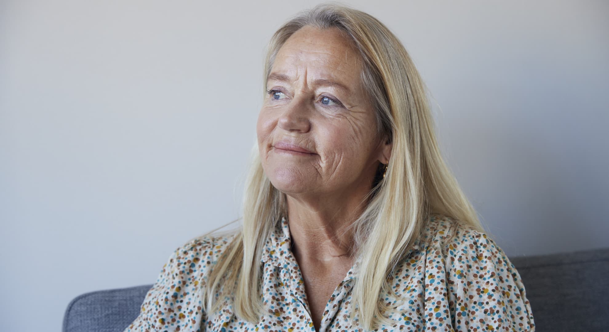Ældre kvinde med langt blond hår og blomstret skjorte ser eftertænksomt til siden, sidder foran en neutral baggrund.