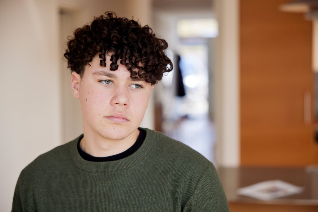 ung person med mørkt, krøllet hår og grøn sweater står indendørs og kigger til siden med et neutralt udtryk.