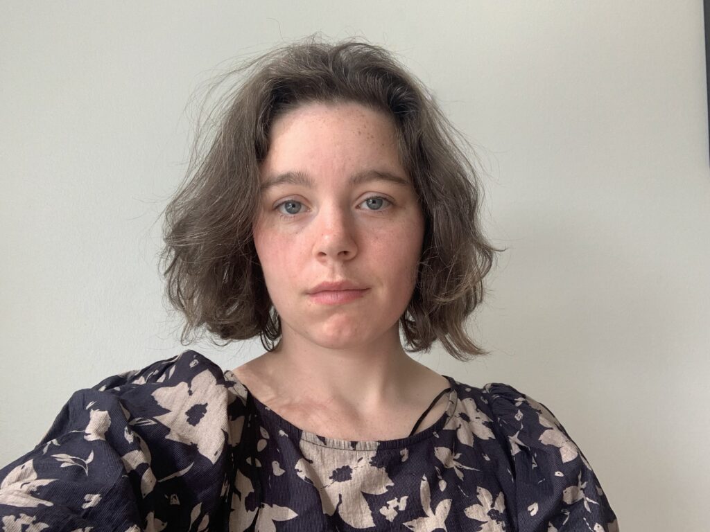 Ung kvinde med kort, brunt hår og blå øjne iført en mørk bluse med blomsterprint, poserer foran en lys baggrund.