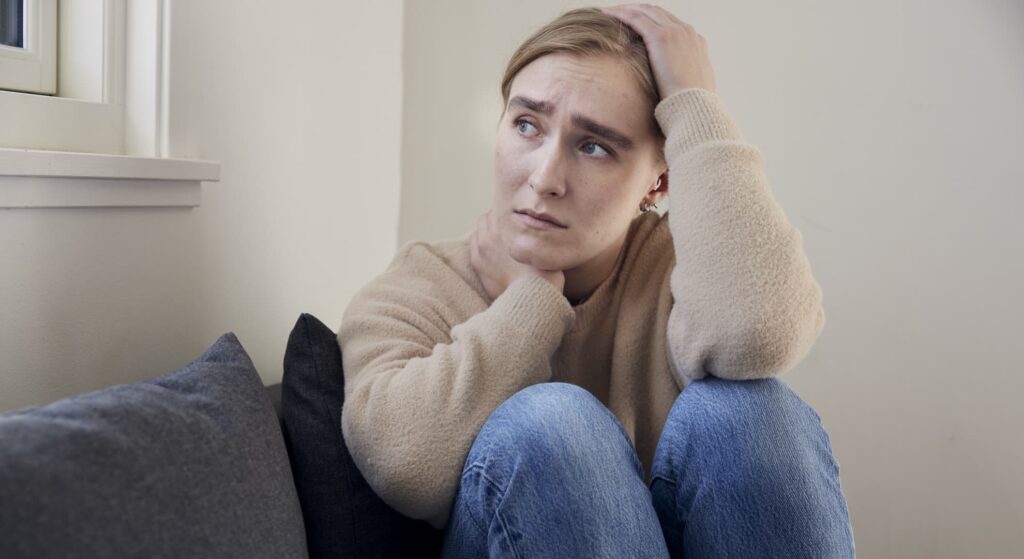 person med bekymret udtryk sidder på en sofa med én hånd i håret og ser til siden.