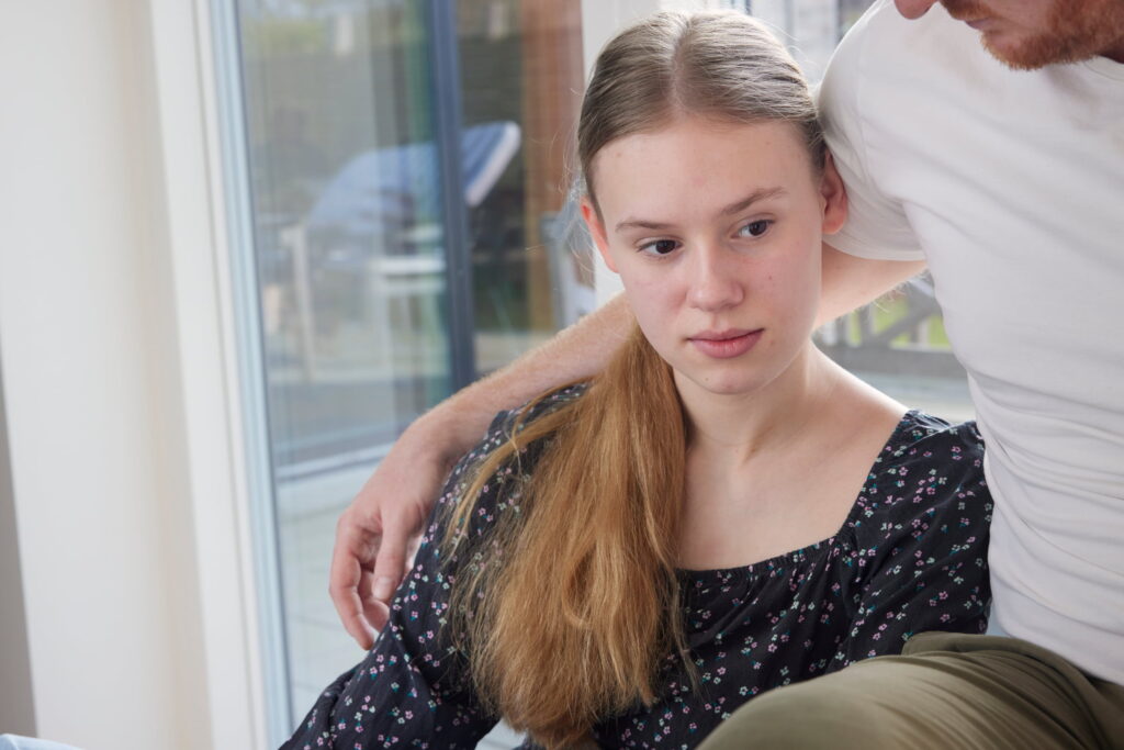 ung kvinde med langt lyst hår sidder indendørs, ser alvorlig ud, mens en person lægger en arm trøstende omkring hendes skuldre.