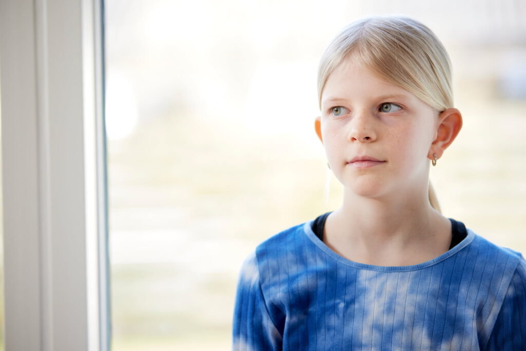 ung pige med lyst hår og blå tie-dye bluse står ved et vindue og kigger ud med et eftertænksomt udtryk.