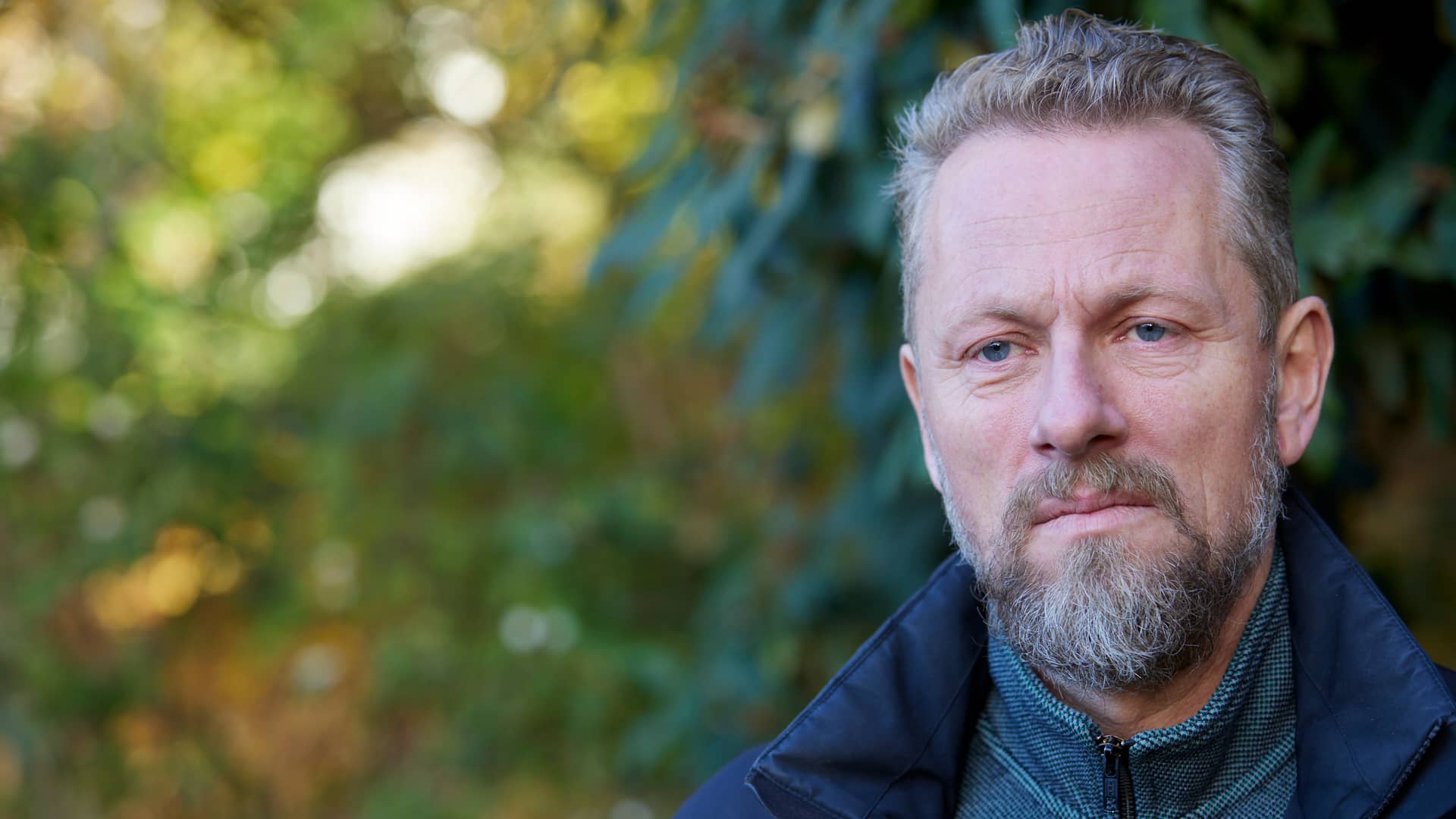 Mand portrætteret udendørs med grønne planter i baggrunden. Han har kort hår, skæg og bærer en mørk jakke. Stemningen er rolig og efterårspræget.