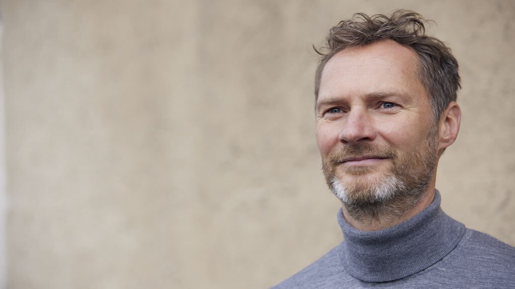 Mand portræt med skæg, iført grå rullekrave, står foran en beige baggrund og smiler let.
