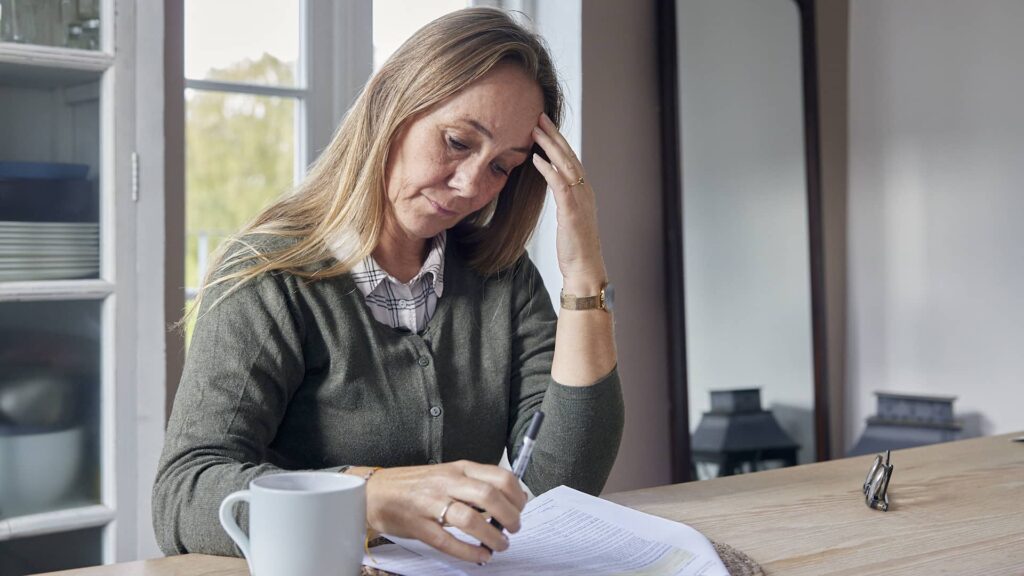 kvinde sidder ved et bord, ser bekymret ud og skriver på nogle dokumenter. En hvid kop står ved siden af hende.