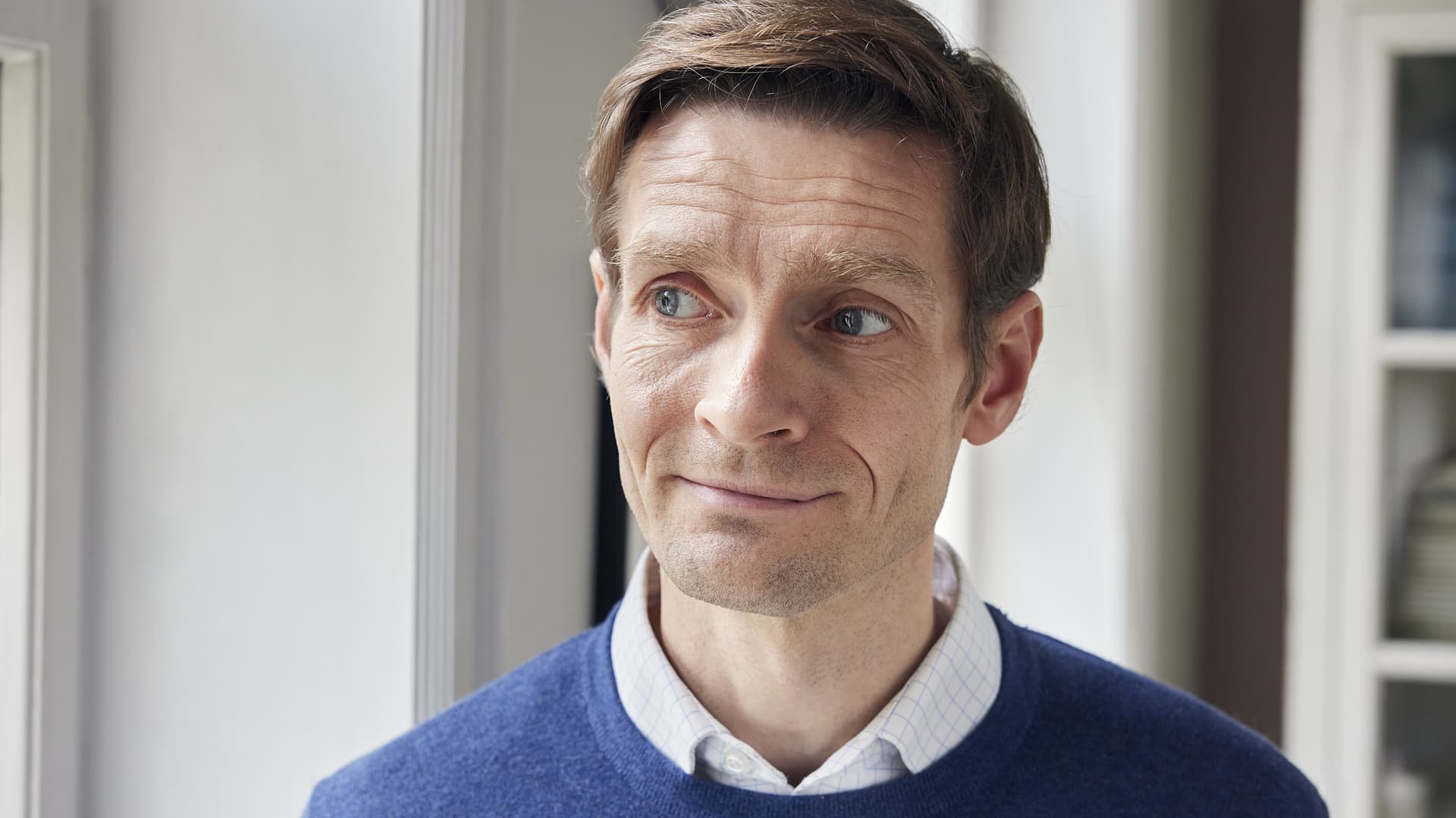 mand med kort brunt hår og blå øjne ser til siden, iført en blå sweater over en hvid skjorte, i et lyst rum.