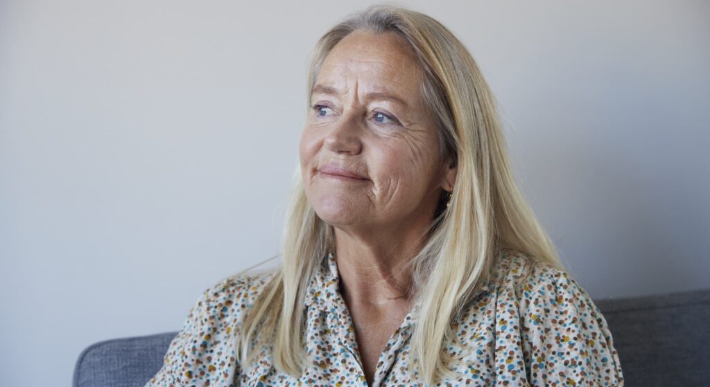Ældre kvinde med langt lyst hår og blomstrede skjorte sidder og smiler let, kigger til siden i et lyst rum.