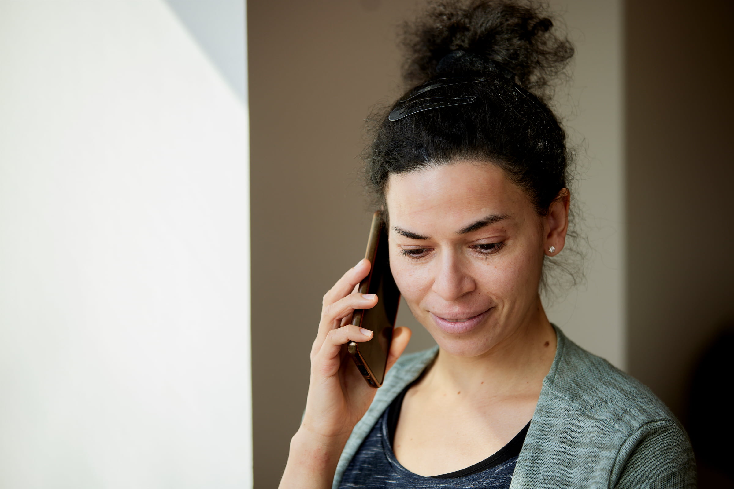 kvinde med mørkt hår taler i telefon og smiler let, mens hun står indendørs i naturligt lys.